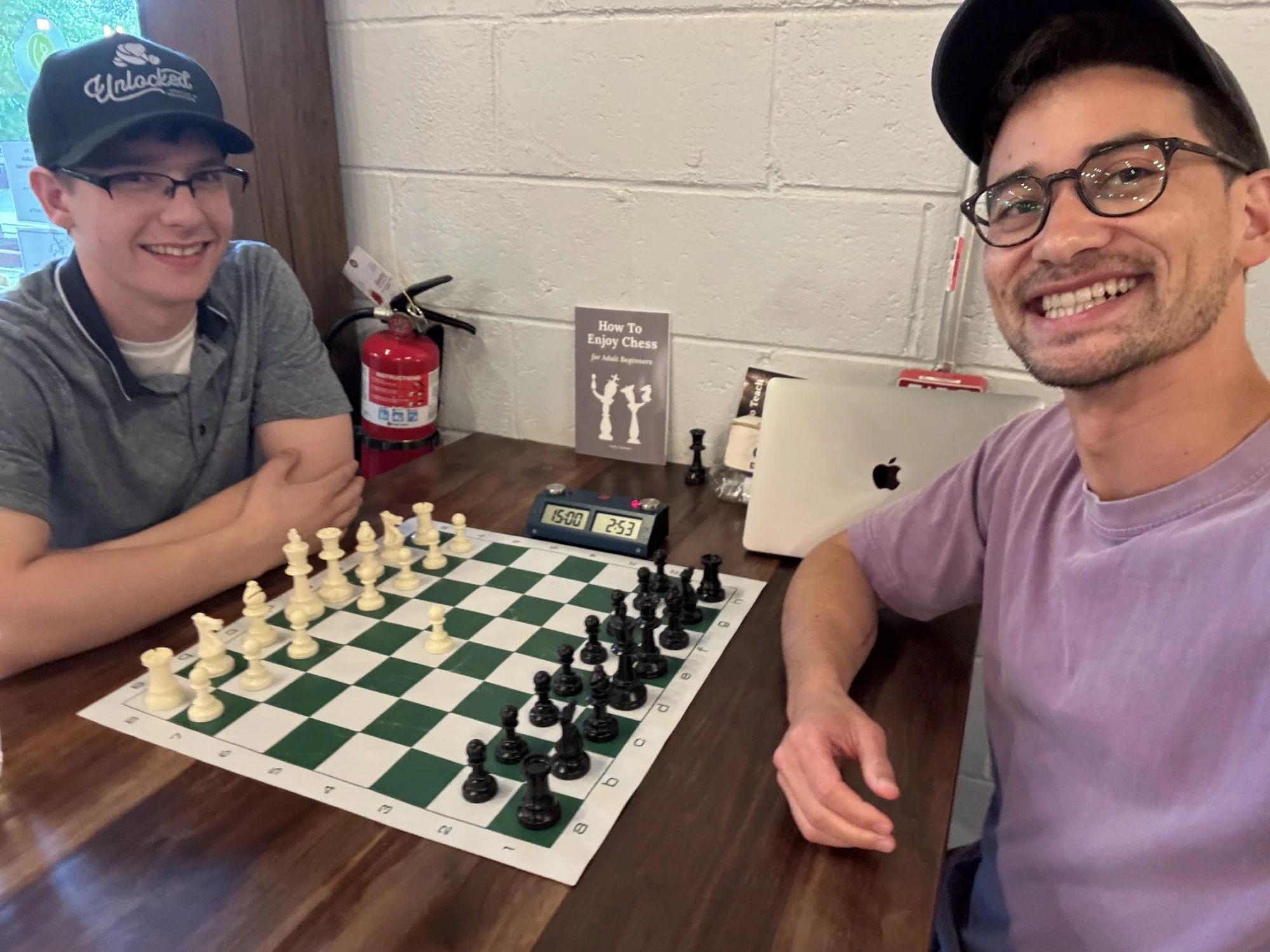 Two people smiling over a chess board mid-game; one wearing an Unlocked cap.